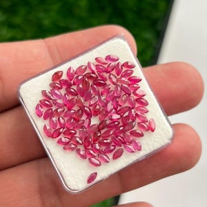 Può includere: Una collezione di piccoli rubini sfaccettati a taglio marquise in una vibrante tonalità rosso rosato. Queste pietre preziose sciolte sono disposte su un inserto in schiuma bianca all'interno di una custodia quadrata in plastica trasparente.