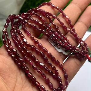 Peut inclure: Un collier de petites perles en pierre précieuse de forme ovale, polies et d'un rouge profond. Les perles sont enfilées sur un fil fin, présentant une qualité translucide et une surface lisse et réfléchissante. Une étiquette blanche avec 7.3.