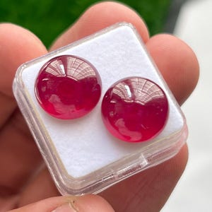 May include: Two round, smooth, deep red cabochon gemstones with a polished, reflective surface. The stones are displayed side-by-side on a white foam insert inside a small, square, clear plastic storage container with a hinged lid.