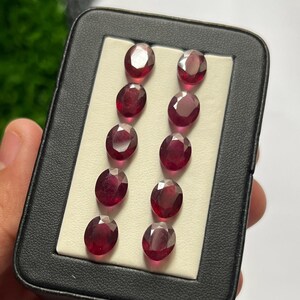 Può includere: Dieci pietre preziose rosso scuro, dal taglio ovale, disposte in due colonne verticali da cinque all'interno di una custodia rettangolare nera. Ogni gemma presenta una superficie sfaccettata che riflette la luce, mostrando un colore cremisi uniforme.