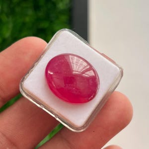 Könnte beinhalten: Ein polierter, oval geformter, leuchtend magentafarbener Edelstein mit einer glatten, reflektierenden Oberfläche. Der Edelstein befindet sich in einer quadratischen, durchsichtigen Kunststoff-Präsentationsbox mit einer weißen Schaumstoffeinlage.