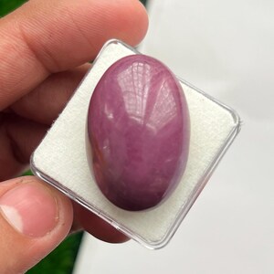 Può includere: Una pietra preziosa lucida di forma ovale con una superficie liscia e brillante di colore rosa magenta intenso. La pietra è inserita all'interno di una custodia espositiva quadrata in plastica trasparente con un inserto in schiuma bianca.