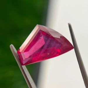 Puede incluir: Una piedra preciosa facetada de forma triangular con un color rojo intenso y vibrante. La piedra presenta múltiples superficies pulidas que reflejan la luz, creando reflejos y sombras internas. Está sujeta de forma segura por dos pinzas metálicas.
