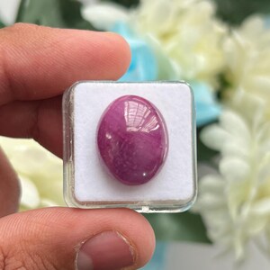 Könnte beinhalten: Ein ovaler, polierter magentafarbener Edelstein mit einer glatten, im Cabochon-Schliff bearbeiteten Oberfläche. Der Stein befindet sich in einer quadratischen, transparenten Kunststoffschatulle mit einer weißen Schaumstoffeinlage.