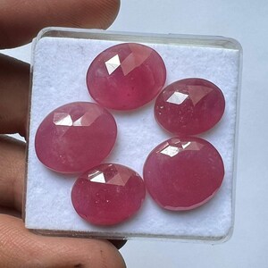 Puede incluir: Cinco gemas facetadas, de forma ovalada, de color rosa-rojo se muestran en un recipiente de plástico transparente. Las gemas tienen una superficie pulida y reflectante con múltiples facetas. La disposición está sobre un fondo blanco.