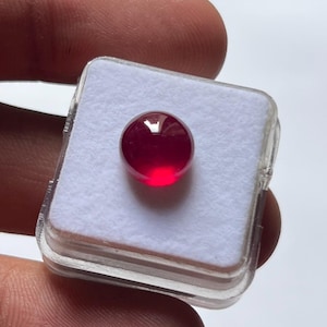 Op de afbeelding: Een ronde, dieprode edelsteen met een glad, gepolijst cabochon-oppervlak. De edelsteen is gecentreerd op een witte schuimrubberen inzet in een klein, vierkant, doorzichtig plastic displaydoosje, wat de levendige kleur en glanzende afwerking benadrukt.