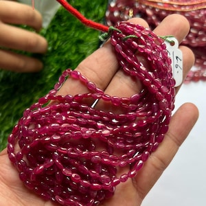 May include: A strand of small, oval-shaped, translucent deep pink beads with a faceted texture. The beads are strung together on a thin green thread, with a white rectangular tag attached that features the handwritten text 6.6.