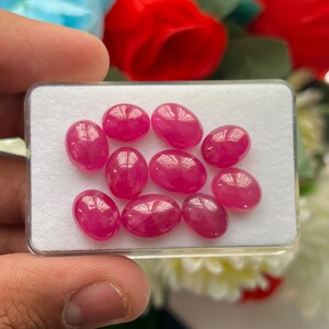 Può includere: Una collezione di nove pietre preziose ovali e levigate di colore rosso rosato, disposte in una scatola espositiva rettangolare bianca con coperchio in plastica trasparente. Ogni pietra presenta una superficie a cabochon liscia e lucida.