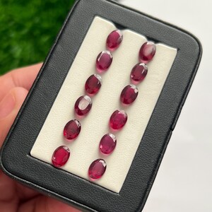 May include: A black rectangular display case containing twelve loose, oval-cut, deep red gemstones arranged in two vertical columns of six on a white foam insert. Each faceted stone exhibits a translucent, vibrant crimson hue and polished finish.
