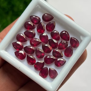 May include: A collection of twenty-two loose, pear-shaped red rubies arranged in a white square display box. Each gemstone features a faceted surface that reflects light, showcasing a deep, saturated crimson color throughout the set.