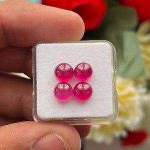 Könnte beinhalten: Vier kleine, runde, durchscheinende fuchsia-pinkfarbene Edelsteine, die in einer quadratischen Formation angeordnet sind. Die Steine liegen auf einer weißen Schaumstoffeinlage in einer quadratischen, transparenten Kunststoffschatulle.