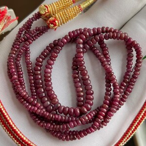 Op de afbeelding: Een ketting met meerdere strengen, samengesteld uit kleine, ronde, dieprode robijnkralen. De ketting heeft goudkleurige accenten bij de sluiting en rood-gouden decoratieve koorden. De kralen zijn aan elkaar geregen, waardoor een gelaagd effect ontstaat.