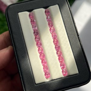 Può includere: Due file verticali di piccole pietre preziose rosa a taglio ovale sono disposte su un inserto in schiuma bianca all'interno di una custodia rettangolare nera. Ogni fila contiene dieci pietre sfaccettate con una tonalità rosa vibrante e traslucida.