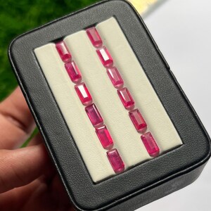 Op de afbeelding: Een verzameling van tien rechthoekige, baguette-geslepen levendige roze edelstenen, gerangschikt in twee parallelle rijen van vijf in een zwarte displaydoos met een witte inzet. Elke edelsteen heeft een gefacetteerd oppervlak en een dieproze kleur.