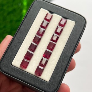 Puede incluir: Un estuche expositor rectangular negro que contiene doce rubíes rojos facetados de corte esmeralda, dispuestos en dos filas verticales paralelas de seis sobre un inserto blanco. Las gemas son de color rojo intenso, translúcidas y geométricas.