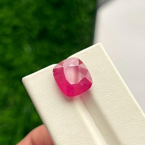Puede incluir: Una piedra preciosa de corte cojín con un tono rosa rojizo vibrante y saturado. La piedra presenta una superficie facetada que refleja la luz, creando una apariencia brillante. Está colocada sobre un soporte de exhibición blanco con una superficie texturizada.