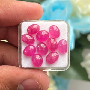 Könnte beinhalten: Eine Ansammlung von zehn kleinen, ovalen, durchscheinenden rosa Edelsteinen, die auf einer weißen Schaumstoffeinlage in einer quadratischen, durchsichtigen Kunststoff-Präsentationsbox angeordnet sind. Jeder Stein hat eine glatte Oberfläche.