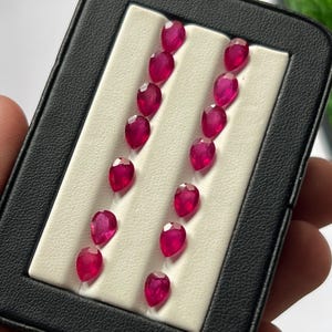 Könnte beinhalten: Eine Sammlung von vierzehn losen, birnenförmigen Rubinen mit leuchtend roter Farbe und facettierten Oberflächen, angeordnet in zwei vertikalen Reihen in einem schwarzen Etui mit einer weißen, strukturierten Einlage zur sicheren Aufbewahrung.