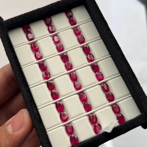 Könnte beinhalten: Eine schwarze Präsentationsbox mit siebenundzwanzig oval geschliffenen, tiefroten Rubinen, die in drei vertikalen Spalten angeordnet sind. Jeder facettierte Edelstein steckt sicher in einem geschlitzten, weißen Schaumstoffeinsatz.