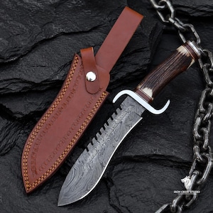 Puede incluir: Un cuchillo de hoja fija con una hoja de acero de Damasco estampada, un lomo dentado y un mango texturizado de color marrón oscuro. El cuchillo cuenta con una guarda de doble gavilán plateada y una funda de cuero marrón con cierre de botón.