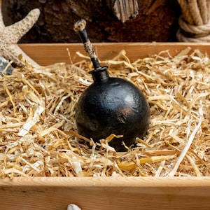 Könnte beinhalten: Eine schwarze, runde Bombenattrappe mit Holzgriff liegt in einer Holzkiste mit Stroh. Ein Seestern und ein Stein im Hintergrund deuten auf ein nautisches oder Piraten-Thema hin.