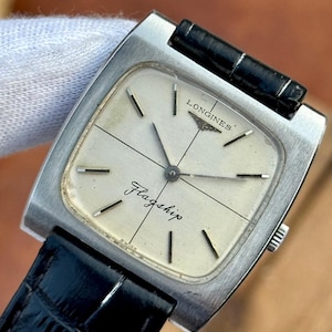May include: A vintage Longines Flagship watch with a silver square case and a cream face. It has black hands and markers, and a black leather strap. The word "Flagship" is printed on the watch face.