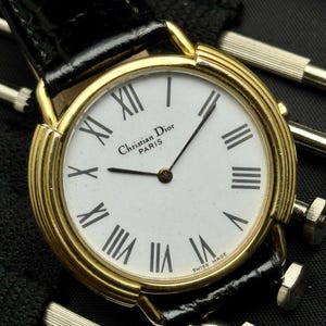 May include: A gold-tone wristwatch with a white dial, black Roman numeral hour markers, and the text Christian Dior PARIS SWISS MADE. The watch features a black leather strap with a textured finish and thin gold-tone lugs on the round case.