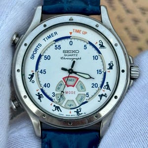 May include: A Seiko Quartz Chronograph watch with a white dial, blue leather strap, and silver-tone case. The dial features a Sports Timer display with various sport icons, a central mode indicator, and the text Seiko Quartz Chronograph.