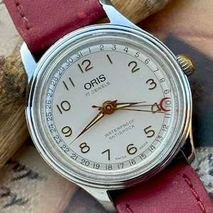 Puede incluir: Un reloj de pulsera Oris plateado con esfera color crema, números arábigos negros, una aguja de fecha con punta de media luna roja y una correa de cuero roja. La esfera muestra el texto Oris, 17 Jewels, Waterproof, Antishock, Swiss Made.