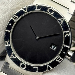 Könnte beinhalten: Eine silberfarbene Armbanduhr mit einem runden schwarzen Zifferblatt, dünnen schwarzen Zeigern und einem Datumsfenster mit der Zahl 23. Die Lünette ist mit dem Schriftzug BVLGARI in Großbuchstaben graviert. Die Uhr hat ein Metallgliederarmband.