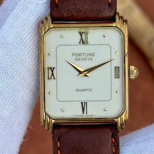 May include: A gold-toned Fortune Genève quartz watch with a rectangular face and brown leather strap. The watch face is white with black Roman numerals and gold hands. The words "Fortune Genève", "Quartz", and "Swiss Made" are visible.