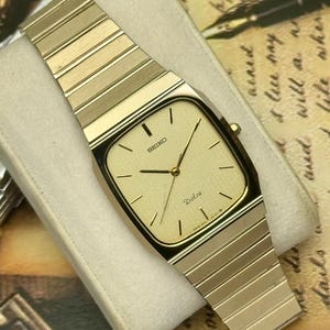 May include: A gold-tone Seiko Dolce wristwatch featuring a square-shaped case with rounded corners and a textured gold dial. The watch includes a matching gold-tone integrated link bracelet, thin black hour markers, and slender gold hands.