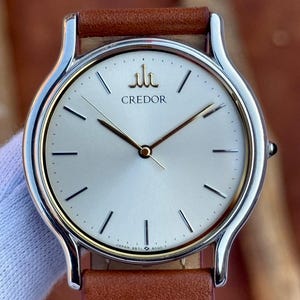 May include: A silver-toned Credor watch with a light-colored face, gold accents, and a brown leather band. The watch face features gold hands and black markers. The band is brown leather with visible stitching. The watch is a classic timepiece.