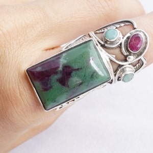 May include: A silver ring with a large green and purple gemstone, surrounded by smaller green and red gemstones.