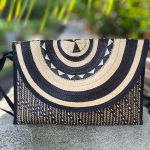 May include: A rectangular woven clutch with a black and natural color pattern. The top flap features concentric semi-circles in black and natural tones, with a triangle at the center. The body of the bag has a woven pattern. A black strap is attached.