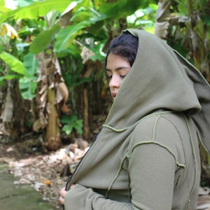 Puede incluir: Una persona lleva una chaqueta verde con capucha y una capucha larga y puntiaguda. La chaqueta tiene una cremallera y está hecha de una tela suave y cómoda. La persona está de pie en un exuberante bosque verde.