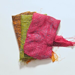 Pode incluir: Três lenços coloridos são exibidos em leque contra um fundo branco. Os lenços são em tons de laranja, verde e rosa. O lenço rosa tem listras prateadas e detalhes azuis. Os lenços têm bordas desfiadas e uma aparência texturizada.