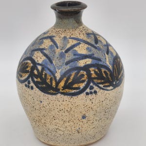 May include: A speckled ceramic vase with a rounded body and a narrow neck. The vase features a decorative band of blue and brown leaf and floral patterns around the middle. The rim is dark brown.
