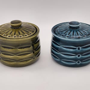 May include: Two ceramic jars with lids. One is olive green, the other is teal blue. Both have a textured design of horizontal lines and small circles. The lids have a sunburst pattern and a small knob.