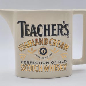 May include: A cream-coloured ceramic jug featuring the black and gold text Teacher's Highland Cream Perfection of Old Scotch Whisky. The vessel has a wide pouring spout, a flat base, and a rectangular handle on the right side of the body.