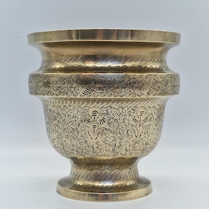 May include: A brass-toned vase featuring detailed floral engravings. The vase has a wide rim, a rounded central section, and a pedestal base. The surface is covered in an elaborate pattern of leaves and flowers.