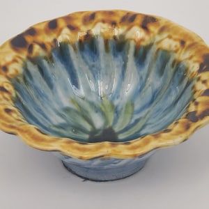 May include: A ceramic bowl with a scalloped rim featuring a brown and tan mottled pattern. The interior of the bowl displays a glossy glaze with streaks of blue, green, and white radiating from the centre towards the textured, wavy edge.