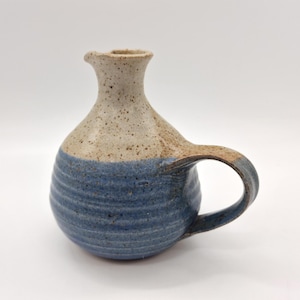 May include: A handmade ceramic jug with a rounded body and a handle. The top portion is a speckled beige, while the bottom is a textured blue. The jug has a small spout and is likely used for pouring liquids.