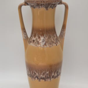 May include: A tall, tan ceramic vase with two handles. The vase has a wide base that tapers upwards, with a brown band and a white, textured band around the middle. The rim is also brown.
