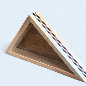 May include: Triangular wooden display case with a natural wood finish. The top edge features three parallel stripes in red, white, and blue. The interior is a darker wood tone, creating a contrast. The case is designed to be mounted on a wall.
