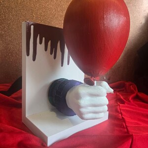 May include: A white L-shaped bookend featuring a sculpted white hand with a dark purple cuff emerging from the centre. The hand holds a glossy red balloon. The vertical back panel of the bookend is decorated with dark red paint dripping from the top edge.