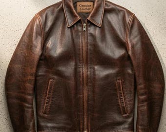 Könnte beinhalten: Braune Lederjacke mit Reißverschluss und zwei Vordertaschen. Die Jacke hat einen Kragen und ein Etikett mit der Aufschrift "Genuine Leather". Das Leder hat ein Used-Look.