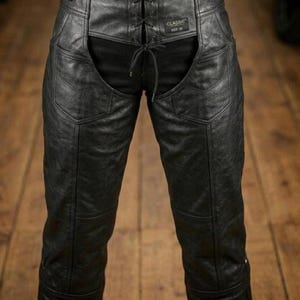 May include: Black leather chaps with a lace-up front and a cutout design. The chaps have a classic style with visible stitching and a small label that reads "CLASSIC SIZE 36". The chaps are shown against a wooden background.