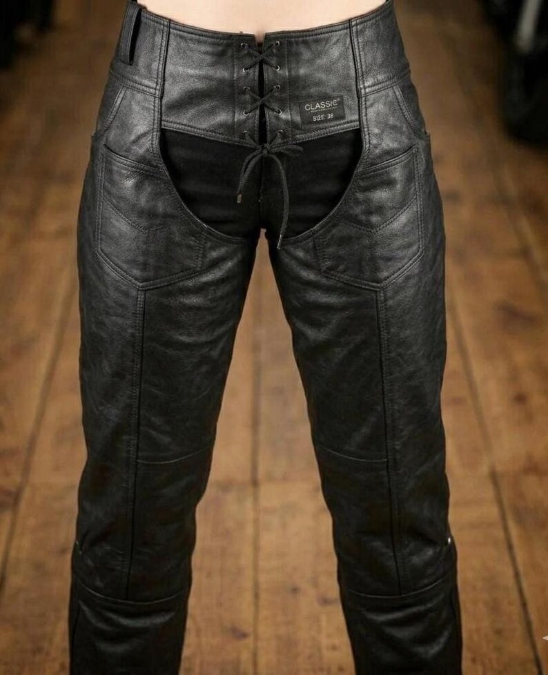 May include: Black leather chaps with a lace-up front and a cutout design. The chaps have a classic style with visible stitching and a small label that reads "CLASSIC SIZE 36". The chaps are shown against a wooden background.