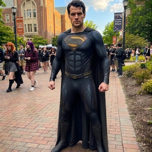 Pode incluir: Um traje completo de Superman preto com um fato texturizado, uma capa longa e um emblema S dourado e preto no peito. O conjunto inclui um cinto a condizer e botas escuras, usado por uma pessoa num caminho de tijolo.